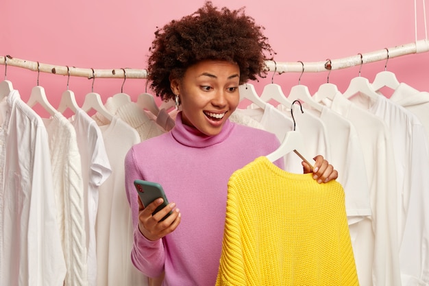 Blij etnische vrouw onderzoekt kledingcollectie in showroom, houdt gele gebreide trui op hanger, maakt gebruik van mobiele telefoon