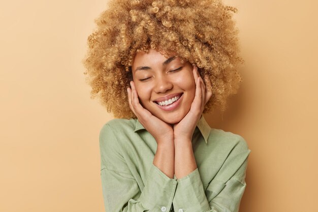 Blij Europese vrouw met krullend haar houdt handen op de wangen glimlacht aangenaam houdt ogen gesloten herinnert zich aangename herinneringen draagt shirt geïsoleerd over beige achtergrond Gelukkig gevoelens concept