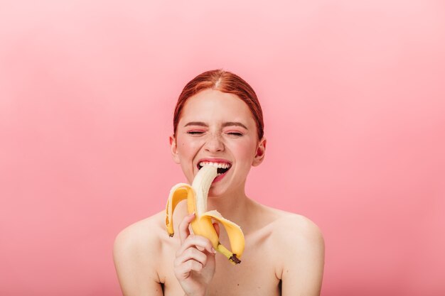 Blij gembermeisje dat banaan eet. Vooraanzicht van tevreden vrouw die van tropisch fruit op roze achtergrond geniet.