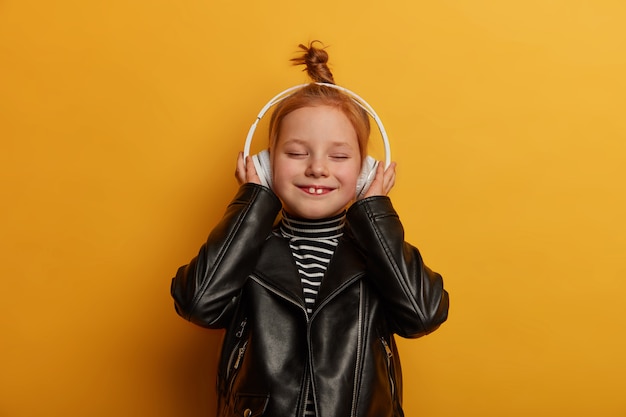 Blij gembermeisje toont twee tanden, luistert naar muziek in een koptelefoon, gekleed in een leren jas, sluit de ogen met plezier, brengt vrije tijd alleen door, geïsoleerd over gele muur. Kinderen, amusement