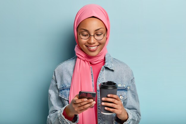 Blij gemengd ras vrouw met gezonde donkere huid, aangename glimlach, houdt mobiele telefoon en afhaalkoffie vast, draagt roze hijab hoofddoek, spijkerjasje, blij om bericht te ontvangen, geïsoleerd over blauwe muur
