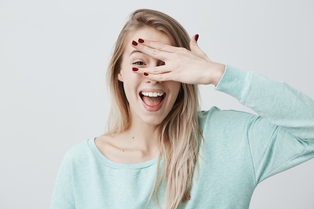 Blij glimlachend blonde vrouwelijk model dat haar gezicht achter hand verbergt, heeft brede glimlach