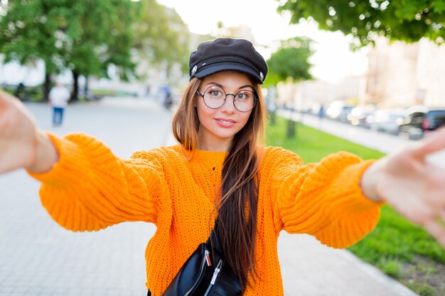 Blij glimlachend donkerbruin meisje in ronde oogglazen en trendy oranje gebreide sweater die zelfportret maken