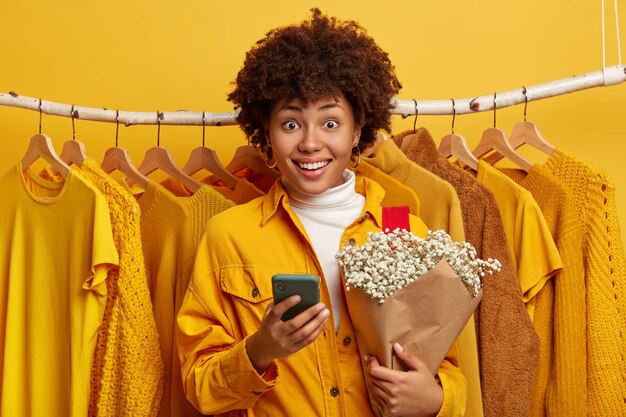 Blij glimlachende Afro-Amerikaanse dame houdt boeket bloemen en moderne mobiele telefoon, vormt in de buurt van kledingrek opknoping op de achtergrond