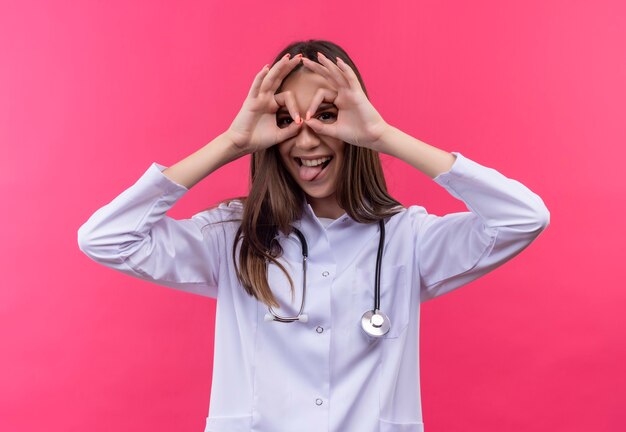 Blij jong artsenmeisje dat stethoscoop medische toga draagt die tong toont en kijkt gebaar op geïsoleerde roze muur