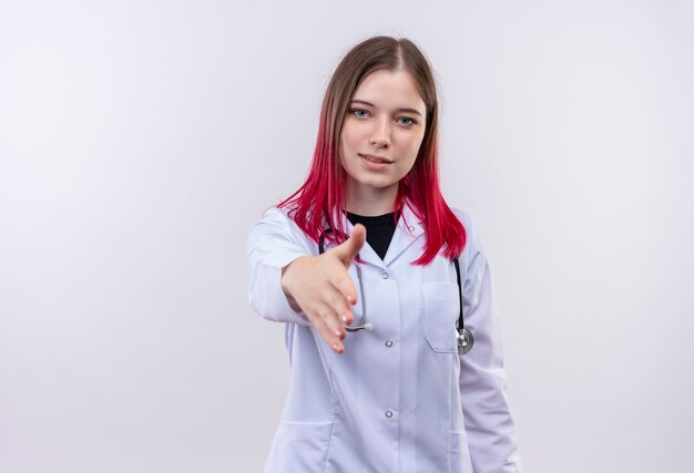 Blij jong artsenmeisje die stethoscoop medisch kleed dragen dat hand uithoudt aan camera op geïsoleerde witte achtergrond