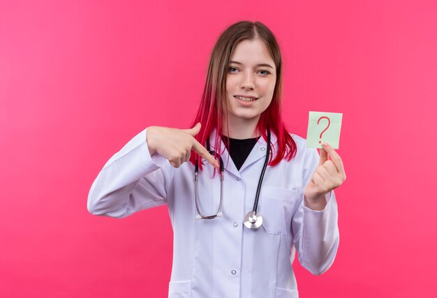 Blij jong artsenmeisje die stethoscoop medische toga dragen wijst naar document vraagteken in haar hand op roze geïsoleerde achtergrond