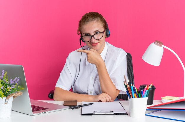 Blij jong blond callcenter meisje met hoofdtelefoon en bril zittend aan een bureau met uitrustingsstukken kijkend naar camera wijzend naar kant geïsoleerd op roze muur