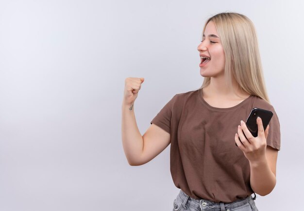 Blij jong blondemeisje die in tandsteunen mobiele telefoon met opgeheven vuist en gesloten ogen op geïsoleerde witte ruimte met exemplaarruimte houden