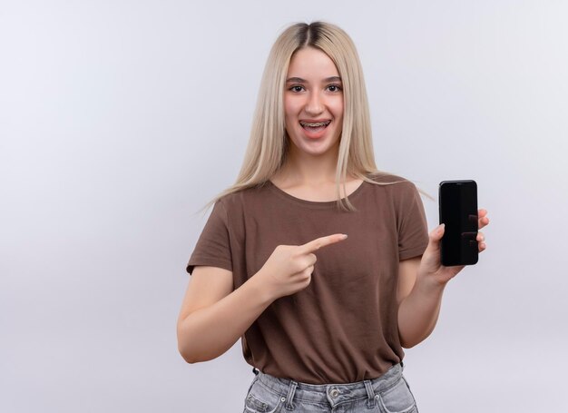 Blij jong blondemeisje in tandsteunen die en op mobiele telefoon op geïsoleerde witte ruimte met exemplaarruimte houden richten
