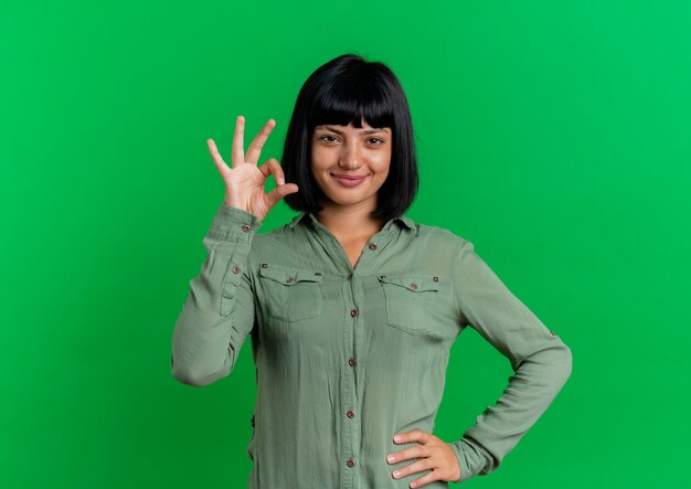 Blij jong brunette Kaukasisch meisje legt hand op taille gebaren ok handteken geïsoleerd op groene achtergrond met kopie ruimte