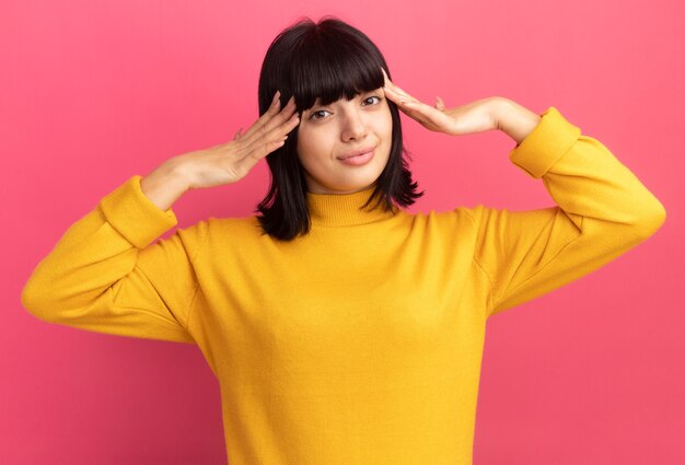 Blij jong brunette kaukasisch meisje legt handen op hoofd geïsoleerd op roze muur met kopieerruimte