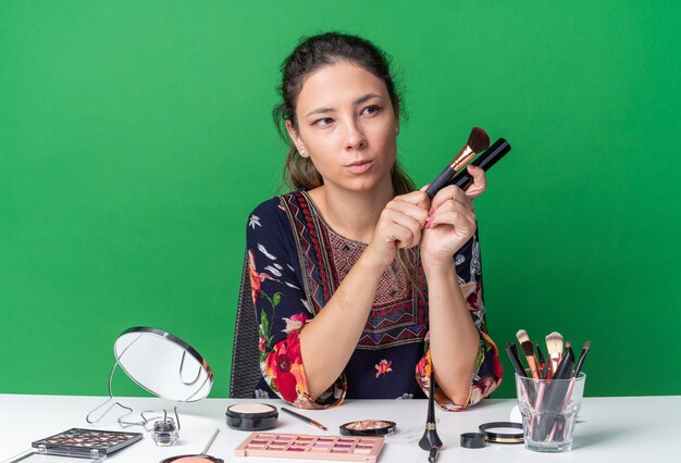 Blij jong brunette meisje zit aan tafel met make-up tools met make-upborstel en mascara die naar de zijkant kijken