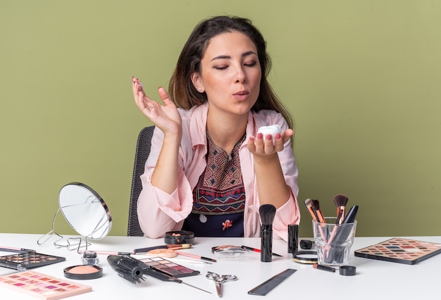Blij jong brunette meisje zittend aan tafel met make-up tools houden en blazen op haar mousse geïsoleerd op olijfgroene muur met kopieerruimte