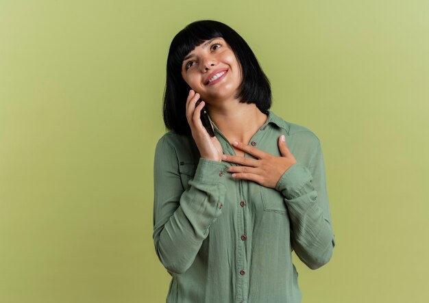 Blij jong donkerbruin Kaukasisch meisje legt hand op borst die op telefoon spreekt en omhoog geïsoleerd op olijfgroene achtergrond met exemplaarruimte kijkt