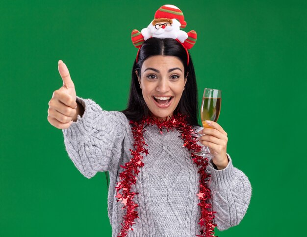 Blij jong Kaukasisch meisje dat de hoofdband van de Kerstman en klatergoudslinger om hals draagt die glas champagne houdt die duim toont die op groene muur wordt geïsoleerd