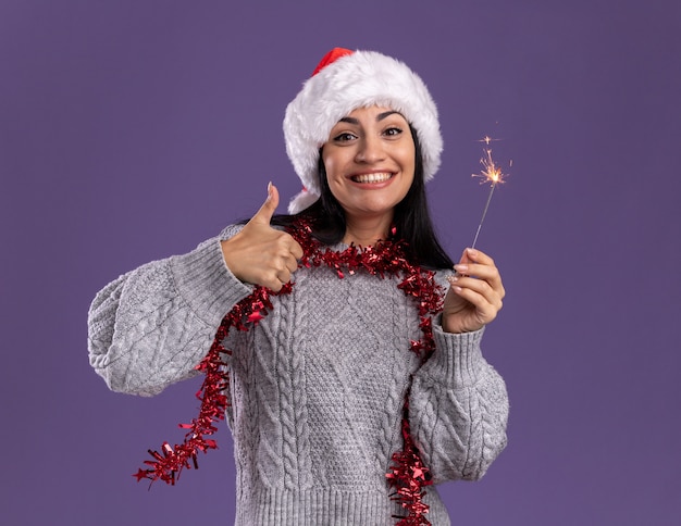 Blij jong kaukasisch meisje dat kerstmishoed en klatergoudslinger om hals draagt die vakantiesterretje houdt dat duim toont omhoog geïsoleerd op purpere muur