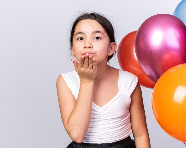 blij jong kaukasisch meisje dat met heliumballonnen staat en kus verzendt met de hand geïsoleerd op een witte muur met kopieerruimte