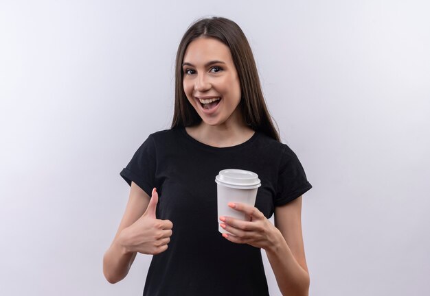 Blij jong Kaukasisch meisje die zwarte t-shirt dragen die kop van koffie haar duim omhoog op geïsoleerde witte muur houden