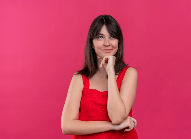 Blij jong Kaukasisch meisje hand op kin zetten en kijken op geïsoleerde roze achtergrond met kopie ruimte