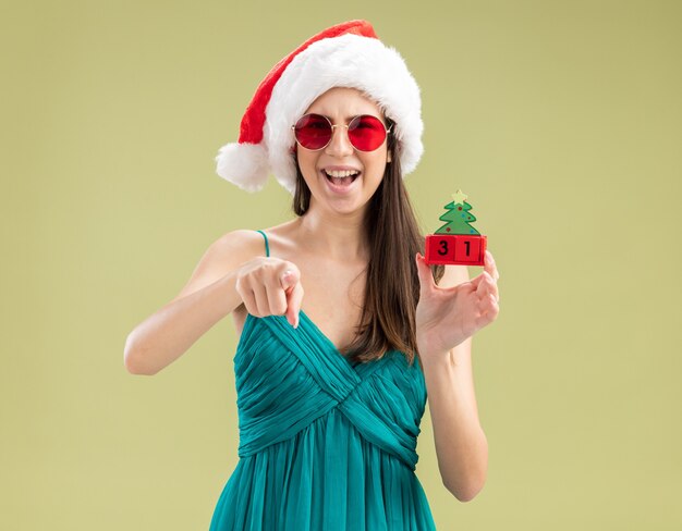 Blij jong Kaukasisch meisje in zonglazen met kerstmuts met kerstboom ornament en wijzen