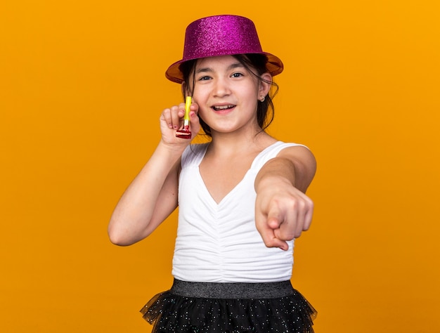 Blij jong kaukasisch meisje met paarse feestmuts partij fluitje houden en wijzen geïsoleerd op oranje muur met kopie ruimte