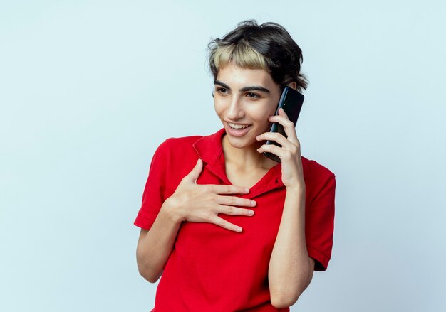 Blij jong Kaukasisch meisje met pixiekapsel dat aan telefoon met hand op borst spreekt die recht kijkt geïsoleerd op witte achtergrond met exemplaarruimte