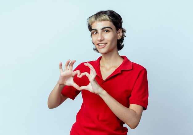 Blij jong Kaukasisch meisje met pixiekapsel die hartteken doen dat op witte achtergrond met exemplaarruimte wordt geïsoleerd