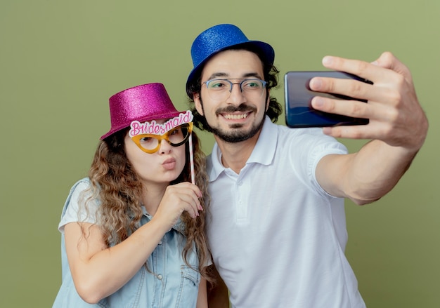 Blij jong koppel met roze en blauwe hoed nemen een selfie en meisje met maskerade oogmasker op stok