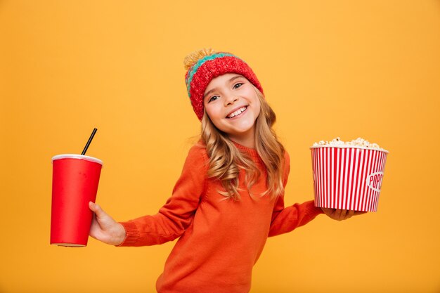 Blij Jong meisje in sweater en hoedenholding popcorn en plastic kop terwijl het bekijken de camera over sinaasappel