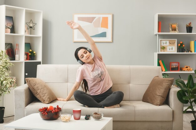 Blij jong meisje met een koptelefoon die yoga doet, zittend op de bank achter de salontafel in de woonkamer