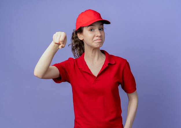 Blij jong mooi bezorgmeisje dat een rood uniform en een pet draagt, doet alsof hij iets vasthoudt dat op paarse achtergrond met kopie ruimte wordt geïsoleerd