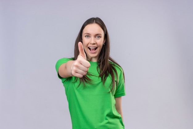 Blij jong mooi meisje dat een groene t-shirt draagt die vrolijk glimlachend duimen toont die zich over geïsoleerde witte achtergrond bevinden