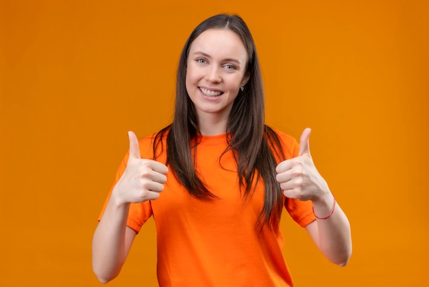 Blij jong mooi meisje dat een oranje t-shirt draagt die vrolijk glimlacht toont duimen met beide handen die zich over geïsoleerde oranje achtergrond bevinden