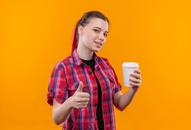 Blij jong mooi meisje dat een rode kop van de overhemdsholding van de koffie haar duim op geïsoleerde gele achtergrond met exemplaarruimte draagt