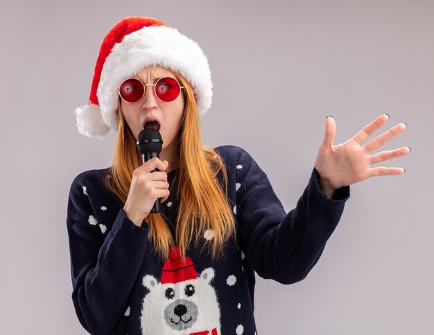 Blij jong mooi meisje met kerstmuts en bril zingen op microfoon spreidende hand geïsoleerd op een witte achtergrond