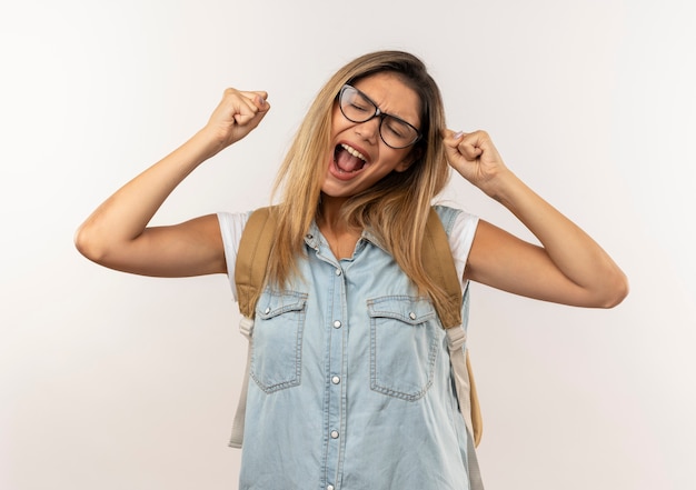 Blij jong mooi studentenmeisje die glazen en achterzak dragen die vuisten met gesloten ogen opheffen die op wit worden geïsoleerd