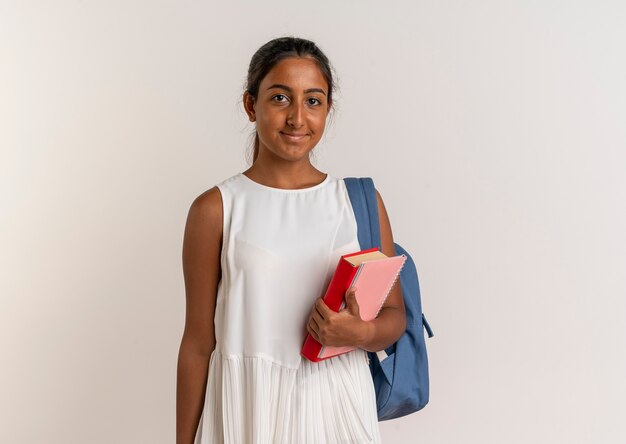 Blij jong schoolmeisje die achterzak met boek met notitieboekje op wit dragen