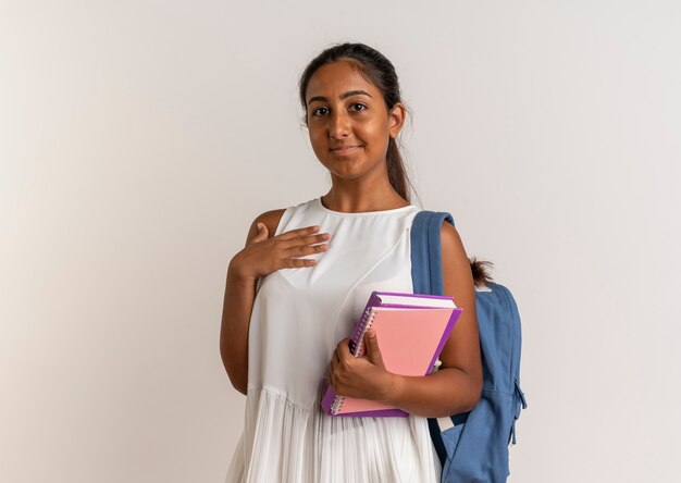 Blij jong schoolmeisje die rugtas dragen die notitieboekje en punten met hand op zichzelf op wit houden