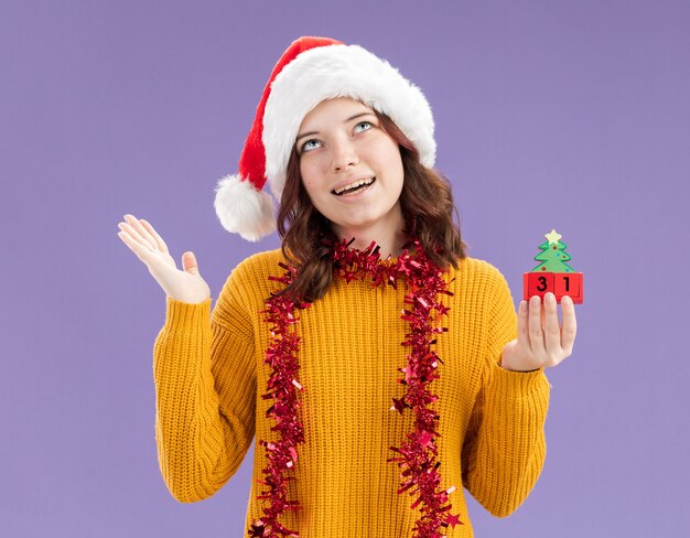 Blij jong Slavisch meisje met kerstmuts en met slinger rond de nek kerstboom ornament te houden en opzoeken geïsoleerd op paarse achtergrond met kopie ruimte