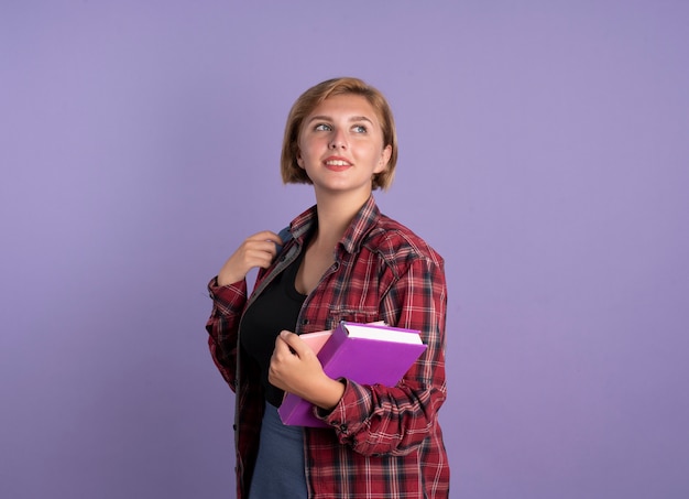 Blij jong slavisch studentenmeisje dat rugzak draagt, staat zijwaarts met boek en notitieboekje