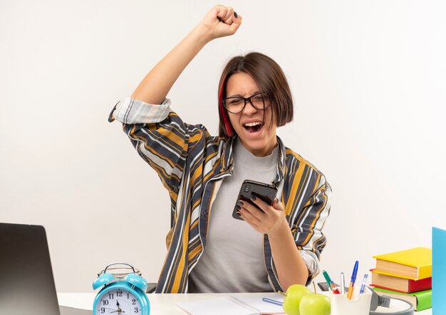 Blij jong studentenmeisje die glazen dragen die bij bureau zitten die mobiele telefoon houden die vuist met gesloten ogen opheffen die op wit worden geïsoleerd