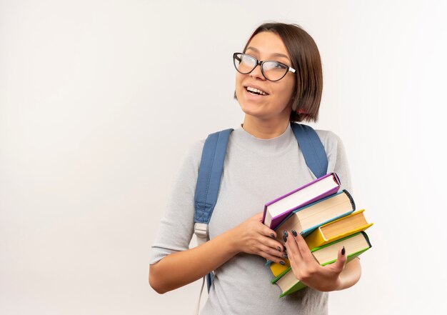 Blij jong studentenmeisje die glazen en de achterboeken van de zakholding dragen die kant bekijken die op wit wordt geïsoleerd