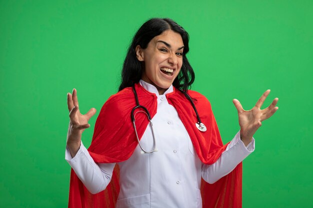 Blij jong superheromeisje die medisch kleed met stethoscoop spreidende handen dragen die op groen worden geïsoleerd