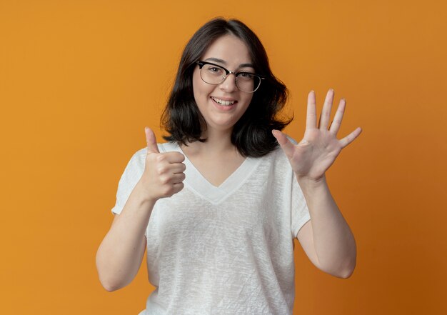 Blij jong vrij Kaukasisch meisje dat glazen draagt die zes met handen tonen die op oranje achtergrond worden geïsoleerd