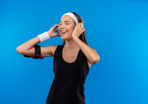 Blij jong vrij sportief meisje die hoofdband en polsbandje en hoofdtelefoons met telefoonarmband dragen die aan muziek met gesloten ogen luisteren