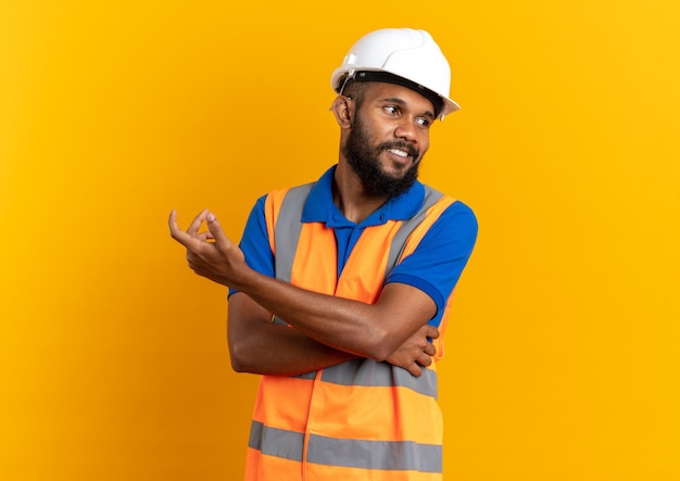 Blij jonge Afro-Amerikaanse bouwer man in uniform met veiligheidshelm kijkend naar kant geïsoleerd op oranje achtergrond met kopie ruimte