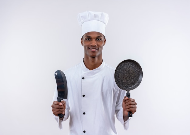 Blij jonge Afro-Amerikaanse kok in uniform chef houdt mes en koekenpan op geïsoleerde witte achtergrond met kopie ruimte
