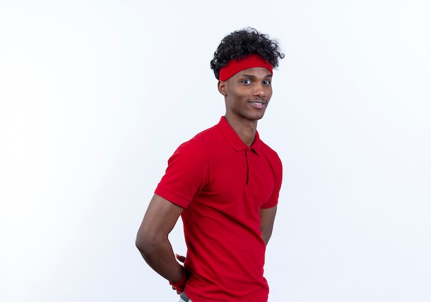 Blij jonge Afro-Amerikaanse sportieve man met hoofdband en polsbandje houden handen achter rug geïsoleerd op een witte achtergrond
