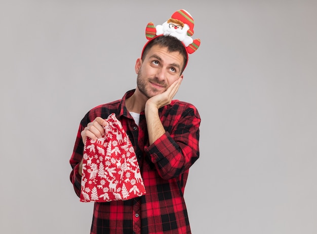 Blij jonge blanke man met kerst hoofdband met kerst zak houden hand op gezicht opzoeken geïsoleerd op een witte achtergrond met kopie ruimte